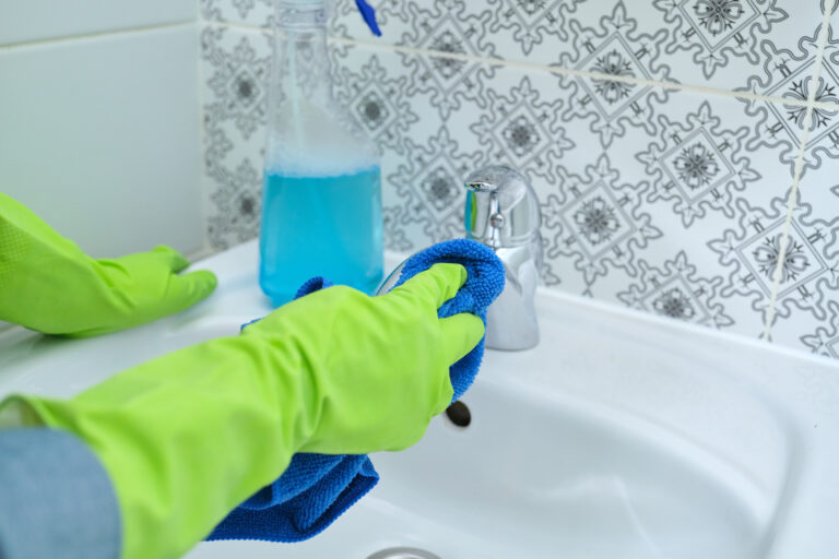 Close up of woman hands in gloves with detergent washing cleaning and polishing wash basin and faucet in bathroom